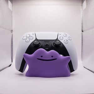 May include: A white and black video game controller with a purple, 3D printed, cartoon character in front of it.