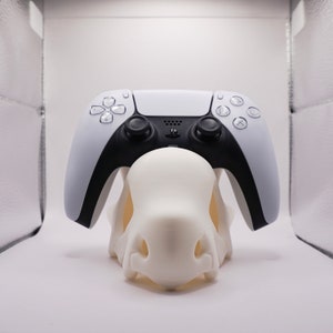 May include: A white PlayStation 5 DualSense controller is sitting on a white 3D printed stand. The controller has black buttons and a black PlayStation logo.