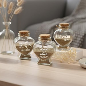 Pode incluir: Garrafas de vidro em forma de coração cheias de areia, seladas com rolhas e personalizadas com nomes e datas. Uma garrafa diz "Hawaii 2025". As garrafas estão dispostas sobre uma mesa de madeira com uma vela, uma suculenta e um vaso de flores secas.