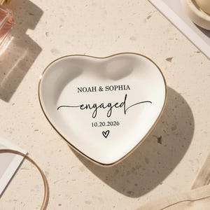 Puede incluir: Un joyero de cerámica blanca en forma de corazón con borde dorado, con la inscripción "Noah & Sophia engaged 10.20.2026" y un símbolo de corazón. También se ven un frasco de perfume, una pulsera dorada y una vela.
