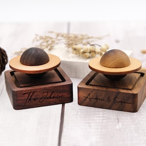 Puede incluir: Dos cajas de anillos de madera con un diseño único. Cada caja tiene una base marrón oscuro con un soporte para anillos marrón más claro. Una caja está grabada con "The Johnson" y la otra con "Andrew & Sophia".