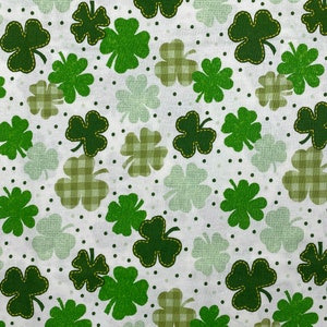 May include: White fabric with a green four-leaf clover pattern. The clover leaves are outlined in yellow thread. The background has small green dots and a checkered pattern.