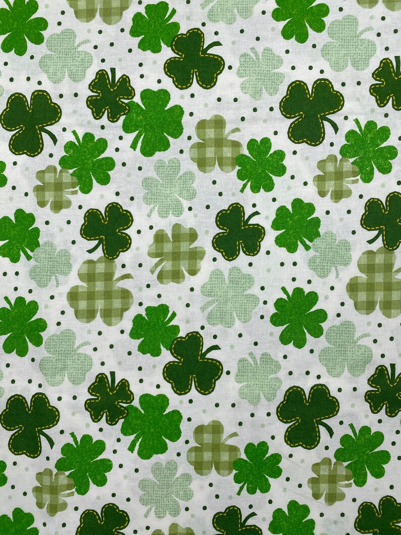 May include: White fabric with a green four-leaf clover pattern. The clover leaves are outlined in yellow thread. The background has small green dots and a checkered pattern.