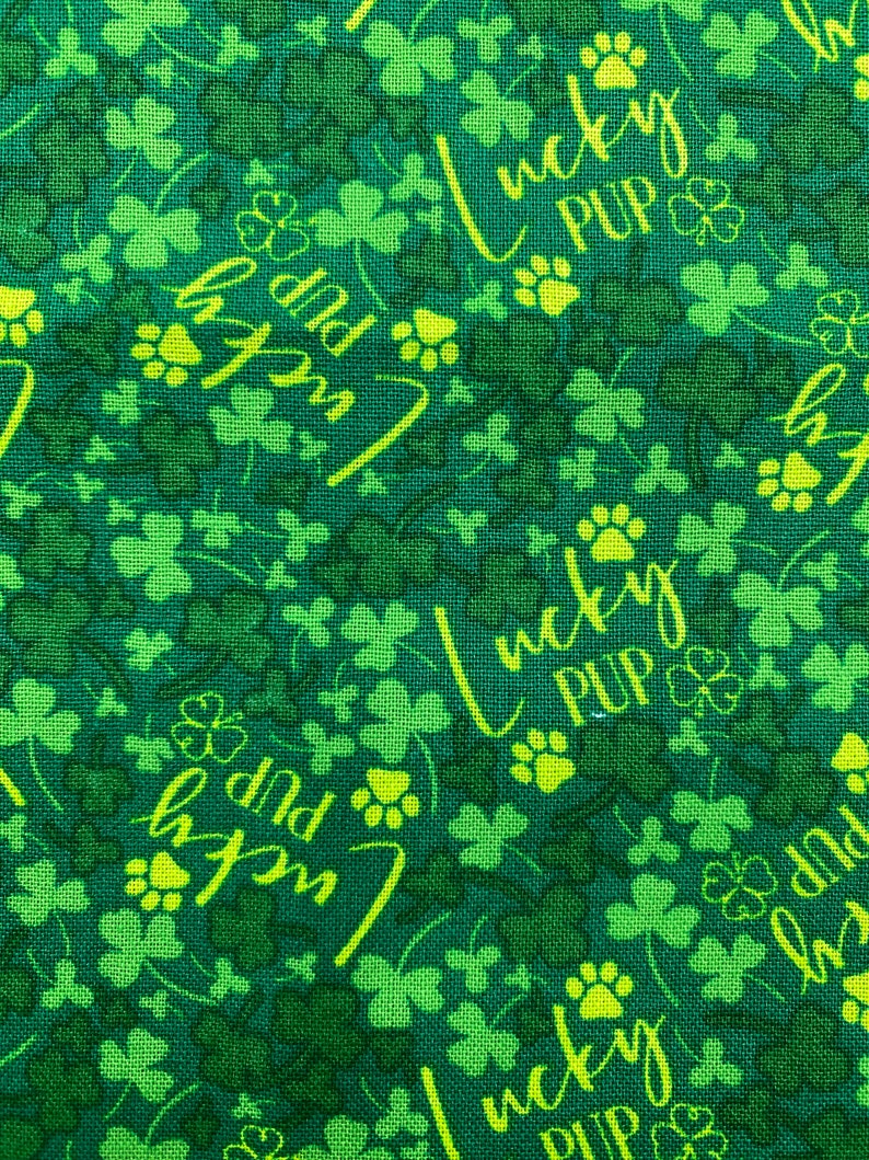 May include: Green fabric with a repeating pattern of shamrocks and paw prints. The text "Lucky Pup" is written in yellow cursive.