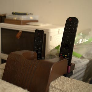 May include: A brown wooden remote control holder with two black remotes. The holder has a curved shape and is designed to hold the remotes upright. The remotes are both standard size and have a black finish.