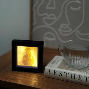 Puede incluir: Un marco de fotos cuadrado negro iluminado que muestra un retrato de graduación. El marco está sobre una superficie de madera, junto a un libro y una taza de cristal transparente. El retrato está iluminado con una luz cálida y amarilla.