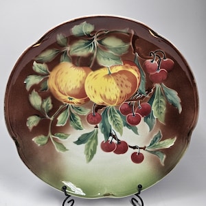 May include: A decorative ceramic plate with a brown background and a painted design of yellow apples and red cherries on branches with green leaves. The plate has a scalloped edge and is displayed on a black metal stand.