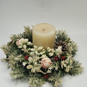 May include: A cylindrical cream-colored pillar candle sits in the center of a circular wreath. The wreath features green and frosted pine needles, white holly leaves, small red berries, a brown pinecone, and two small red and white striped apples.