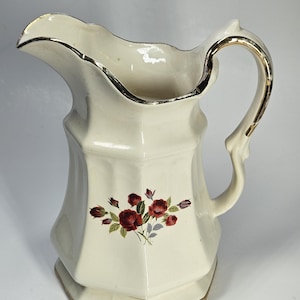 May include: Cream-colored ceramic pitcher with a floral design of red roses and green leaves. The pitcher has a gold trim around the rim, handle, and base. The pitcher has a unique, faceted shape.