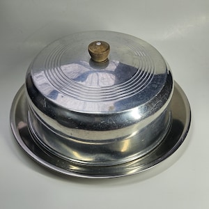 May include: A vintage, silver-colored metal cake carrier with a domed lid and a wooden knob. The carrier sits on a matching round tray. The lid has concentric circular ridges. The cake carrier is designed to protect and transport baked goods.
