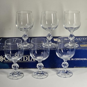 May include: Six clear crystal wine glasses. Each glass has a round bowl, a decorative stem with a faceted design, and a circular base. The glasses are arranged on a white surface with a blue box in the background.