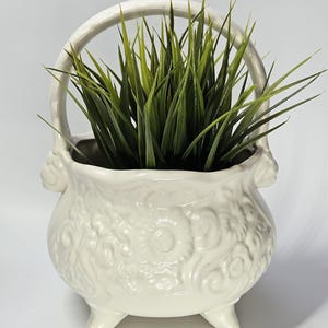 May include: A white ceramic basket-style planter with a handle and three small feet. The planter is filled with green, grass-like plants. The exterior of the planter features a floral embossed design.