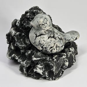 May include: A decorative bird figurine perched on a textured, black and white rock formation. The bird is primarily white with black accents, and the rock has a rough, layered appearance. The sculpture is set against a plain white background.