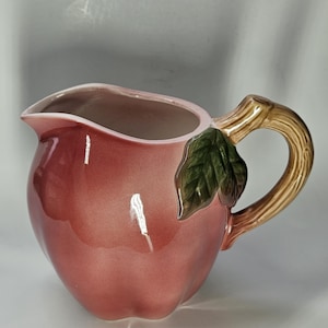 May include: A ceramic pitcher shaped like a red apple with a brown handle and a green leaf accent. The pitcher has a glossy finish and a wide opening for pouring. The handle is designed to resemble a tree branch.