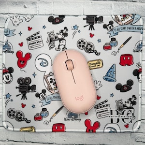 May include: A light pink wireless computer mouse sits on a mousepad with a Disney theme. The mousepad features Mickey Mouse, film reels, and the text "It All Started With a Mouse". The mousepad is rectangular and has a white border.