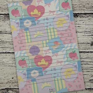May include: A rectangular notepad featuring a patchwork pattern of pastel colours including pink, blue, and yellow. The design includes illustrations of hearts, crowns, apples, seashells, and bows. Horizontal black lines are printed across the paper.