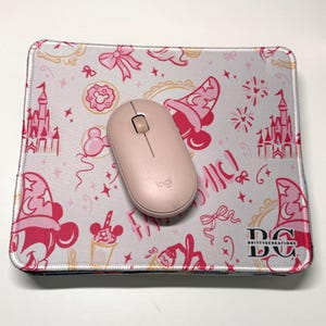 May include: A rectangular mouse pad with a light gray background featuring a pink pattern of castles, wizard hats, bows, and donuts, with a light pink wireless computer mouse centered on top. The logo BC Brittyscreations is printed in black.