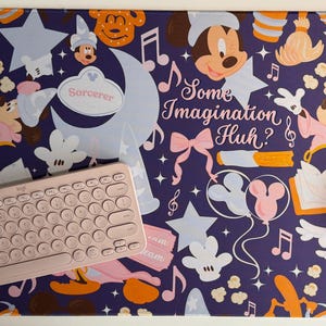 May include: A rectangular desk mat with a dark purple background featuring a repeating pattern of Mickey Mouse, stars, musical notes, and the text "Some Imagination Huh?" and "Sorcerer". A light pink wireless keyboard and mouse sit on the mat.