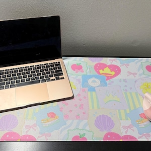 May include: A rose gold laptop and a light pink ergonomic vertical mouse sit on a rectangular desk mat featuring a repeating pattern of pastel hearts, crowns, seashells, and apples in shades of pink, yellow, blue, and white on a light background.