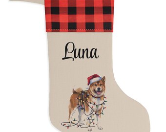 May include: A beige Christmas stocking featuring a red and black buffalo plaid cuff. The name Luna is printed in black script above an illustration of a Shiba Inu dog wearing a Santa hat and tangled in colourful Christmas string lights.