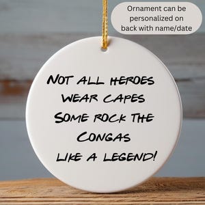 May include: White ceramic ornament featuring the text "Not all heroes wear capes some rock the congas like a legend!" The ornament can be personalised on the back with a name or date.