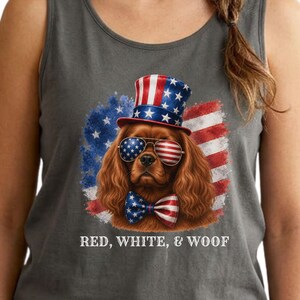 May include: Grey vest top with a Cavalier King Charles Spaniel wearing patriotic sunglasses, a bow tie, and a top hat. The dog is set against a backdrop of the American flag and a blue star field. The text "RED, WHITE, & WOOF" is printed below.