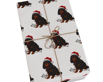 May include: A rectangular gift wrapped in white paper featuring a repeating pattern of black and tan Cavalier King Charles Spaniel dogs wearing red Santa hats and tangled in colorful Christmas string lights, tied with a thin brown twine bow.