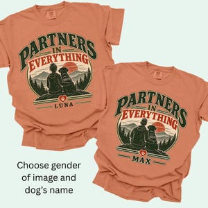 May include: Two terracotta-colored t-shirts featuring a graphic of a person and a dog sitting before mountains and a sun. The text reads PARTNERS IN EVERYTHING with the names LUNA or MAX below a heart icon. The shirts have a crew neck and short sleeves.