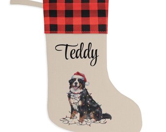 May include: A beige Christmas stocking featuring a red and black buffalo plaid cuff and a hanging loop. The stocking is personalized with the name Teddy in black script and displays a Bernese Mountain Dog wearing a Santa hat and Christmas lights.
