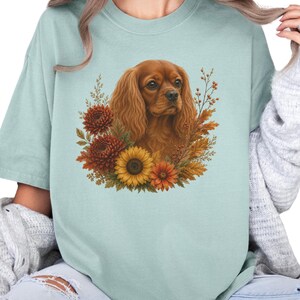 May include: A light sage green t-shirt featuring a central graphic of a brown Cavalier King Charles Spaniel surrounded by a cluster of autumn-coloured flowers, including a large yellow sunflower, dark orange chrysanthemums, and dried foliage.