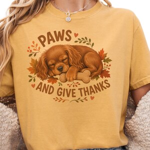 May include: A mustard yellow t-shirt featuring a graphic of a sleeping brown spaniel puppy resting its head on a bone. The text reads PAWS AND GIVE THANKS, surrounded by autumn-colored leaves, small branches, and tiny hearts in a circular arrangement.