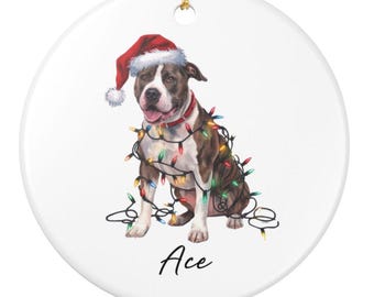 May include: A round, white ceramic ornament featuring a brown and white dog wearing a Santa hat and a red collar, wrapped in colorful Christmas lights. The dog's name, "Ace," is written in black script below.
