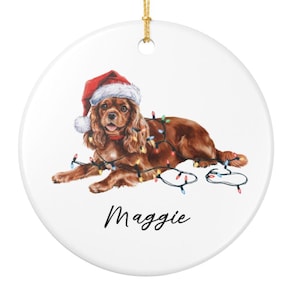 May include: A round, white Christmas ornament featuring a Cavalier King Charles Spaniel wearing a Santa hat and Christmas lights. The dog's name, "Maggie", is written in cursive below the dog.