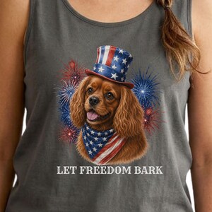 May include: Grey vest top with a Cavalier King Charles Spaniel wearing a patriotic top hat and bandana, with fireworks. The text "LET FREEDOM BARK" is printed below the dog.