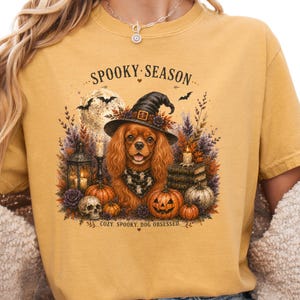 May include: A mustard yellow t-shirt featuring a graphic of a Cavalier King Charles Spaniel wearing a witch hat and a ghost-patterned bandana. The design includes pumpkins, a skull, candles, books, and the text SPOOKY SEASON and COZY. SPOOKY. DOG OBSESSED.