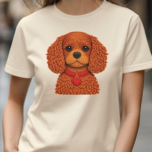 May include: A beige t-shirt featuring a detailed illustration of a brown dog with a red collar and heart-shaped pendant. The dog has large, expressive eyes and a textured fur appearance. The shirt is a casual, everyday wear item.