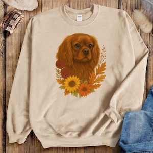 May include: A beige sweatshirt with a detailed illustration of a Cavalier King Charles Spaniel dog, framed by autumn flowers and leaves in orange, yellow, and brown. The dog has long, wavy, reddish-brown fur.