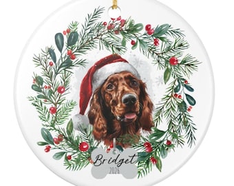 May include: A round white ceramic ornament featuring a brown Irish Setter dog wearing a red and white Santa hat, centered within a green pine wreath with red berries. A bone-shaped tag at the bottom displays the name Bridget and the year 2026.