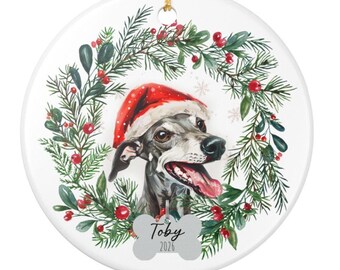 May include: A round white ceramic ornament featuring a watercolor illustration of a grey dog wearing a red Santa hat, centered within a green pine wreath with red berries. A bone-shaped tag at the bottom displays the text Toby and 2026.
