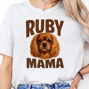 Pode incluir: Camiseta cinza claro com o texto "RUBY MAMA" em letras marrons. A camiseta apresenta uma ilustração detalhada de um cão Cavalier King Charles Spaniel com pelo marrom. O cão está centralizado entre as palavras.
