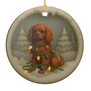 May include: A round ceramic ornament featuring a brown Cavalier King Charles Spaniel wrapped in a string of colorful Christmas lights. The dog is set against a soft, muted background of snow-covered pine trees, with a gold hanging cord at the top.