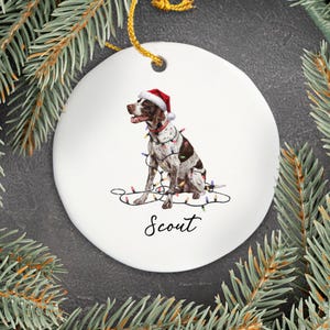 May include: A white ceramic Christmas ornament featuring a brown and white dog wearing a Santa hat and wrapped in Christmas lights. The dog's name, "Scout," is written in black script below the dog. A gold string is attached for hanging.