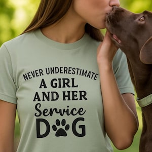 May include: Sage green t-shirt with the text "NEVER UNDERESTIMATE A GIRL AND HER Service DOG" in black. The design includes a paw print graphic. The shirt is made of a soft material.