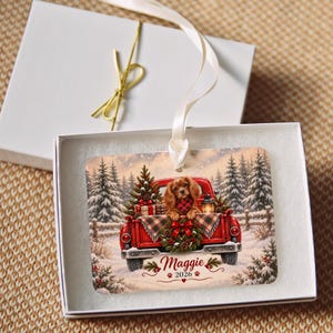 May include: A rectangular Christmas ornament featuring a red pickup truck in a snowy forest with a brown dog wearing a red and black plaid scarf. The truck bed holds a Christmas tree and gifts. The text reads Maggie 2026 with paw prints.