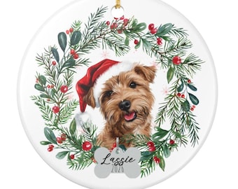 May include: A round white ceramic ornament featuring a brown terrier wearing a red Santa hat, framed by a green pine wreath with red berries. A bone-shaped tag hangs at the bottom with the text Lassie and 2026 printed in black ink.