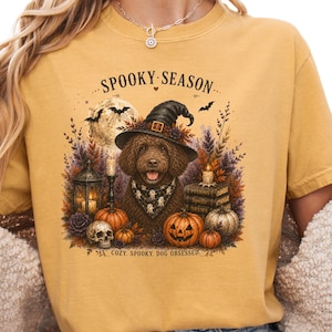 May include: A mustard yellow t-shirt featuring a graphic of a brown dog wearing a witch hat and a ghost-patterned bandana. The design includes a full moon, bats, a lantern, a skull, pumpkins, and books with the text SPOOKY SEASON and COZY. SPOOKY. DOG OBSESSED.