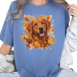May include: A blue t-shirt featuring a graphic print of a golden-colored dog surrounded by vibrant orange and yellow autumn leaves. The dog has a friendly expression with its mouth open, and the design is centered on the chest of the garment.
