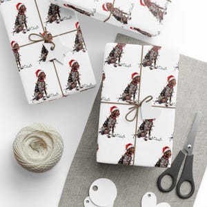 May include: Christmas-themed wrapping paper featuring a repeating pattern of a dog wearing a Santa hat and string lights. The wrapped gifts are tied with twine and a star-shaped tag. A ball of twine, scissors, and gift tags are also present.