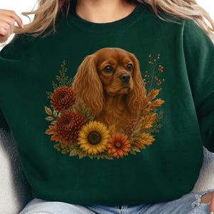 May include: A dark forest green crewneck sweatshirt featuring a central graphic of a brown Cavalier King Charles Spaniel surrounded by a vibrant autumn-themed wreath of orange chrysanthemums, a yellow sunflower, and dried fall foliage.
