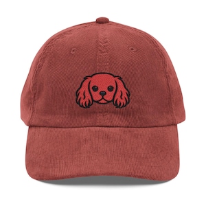 May include: A brick-red corduroy baseball cap featuring an embroidered red Cavalier King Charles Spaniel dog face with black outlines. The cap has a curved brim and an adjustable strap at the back.